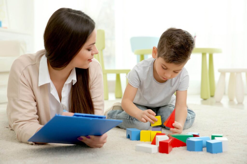 A young woman observes and takes notes as a young boy playing with his blocks