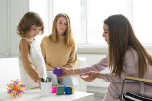 A therapist working with a young child using colorful toys and containers during a therapy session, while the child's guardian observes attentively in a bright, welcoming room.